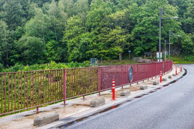 Empty rural road passing over Robertville Dam wall, leafy trees with green foliage in misty blurred background, cloudy day in Waimes, Belgium clipart