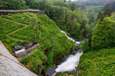Robertville Barajı 'ndan yüksek basınçlı su, yamaçtaki ve küçük binalardaki merdivenler, bulanık ve sisli arka planda yapraklı ağaçlarla dolu Warche vadisi, Waimes, Belçika' da bulutlu bir gün.