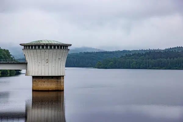 Stock image Gileppe Dam with an intake or outlet tower protruding from lake waters with its bridge, lush green trees in valley mountains in background misty and blurry, cloudy day in Jalhay, Belgium