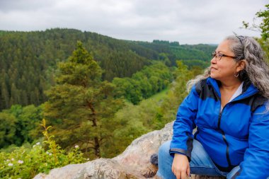 Profilde kıdemli yetişkin kadın yürüyüşçü gülümseyerek manzaraya hayran kalıyor, dağlara bakan vadide kayaları yanıltıyor, arka planda yapraklı ağaçlar, Malmedy, Belçika 'da bulutlu bir gün.