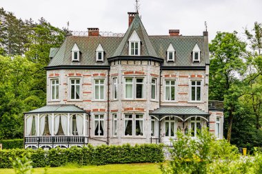 Eski Viktorya tarzı bir evin cephesi, beyaz duvarlar, gri gable çatılar, birden fazla pencere, kapalı veranda ve bahçe, arka planda yeşil yapraklı ağaçlar, Ferrieres, Belçika 'da bulutlu bir gün