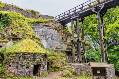 Logne Kalesi 'ndeki bir kaya oluşumunun yanında eski, yuvarlak taştan bir bina, yüksek tahta yaya köprüsü, bol miktarda yosun ve bitki örtüsü, Ferrieres, Belçika' da bulutlu bir gün.
