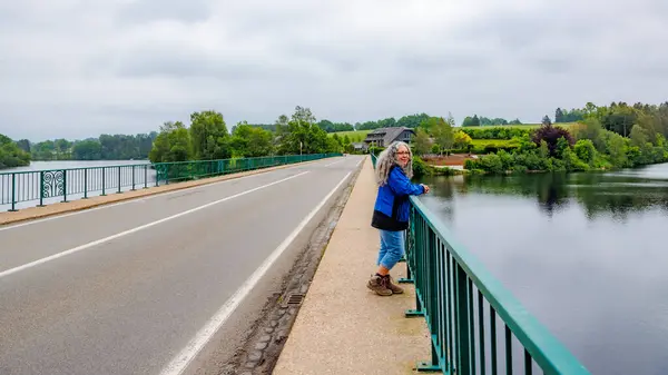 Stock image Vehicular bridge with pedestrian sidewalk over Lake Robertville, older adult woman standing leaning on railing admiring landscape, hills with green trees in background, cloudy day in Waimes, Belgium