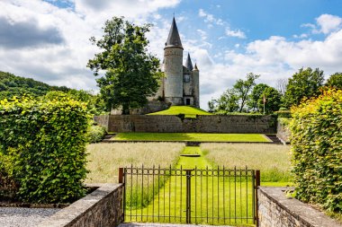 Mavi gökyüzüne karşı tepede metal kapı, bahçeler ve Veves Kalesi, konik çatıları ve taş duvarları olan kuleler, yeşil yapraklı dev ağaç, Houyet, Namur, Wallonia, Belçika 'da güneşli bir gün