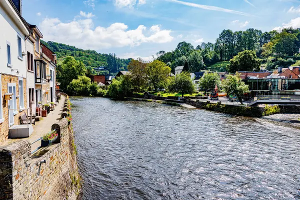 La Roche-en-Ardenne köyündeki Ourthe Nehri, taş duvarlar, evler, binalar ve nehir kıyısı boyunca yayalar, sisli arka planda yeşil ağaçlı dağlar, Belçika 'da güneşli bir yaz günü.