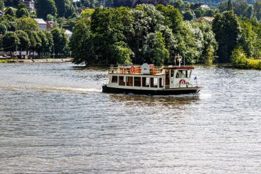 Meuse nehri sakin sularda yüzen boş bir turist teknesi, Dinant şehrinin dışında, arka planda bol yeşil yapraklı ağaçlar, Namur, Wallonia, Belçika 'da güneşli bir yaz günü.