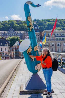 Dinant, Namur, Wallonia, Belçika. 5 Haziran 2024. Dünya haritalı boyanmış saksafon heykelinin yanında poz veren kadın turist bulanık arka plandaki binalar, Adolphe Sax anıtı, güneşli yaz günü