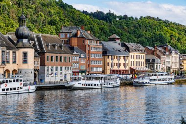 Meuse nehrinin kıyısına demirlemiş turist tekneleri, arka planda yeşil yapraklı ağaçlarla kaplı şehir manzaralı binalar, Wallonia, Belçika 'da güneşli bir yaz günü.