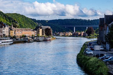 Dinant şehrindeki Meuse nehrinin havadan görünüşü, kıyıya demir atmış tekneler, binalarla dolu şehir manzarası ve arka planda yapraklı ağaçlarla kaplı dağ üzerindeki araç köprüsü, Namur, Wallonia, Belçika