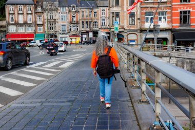 Meuse nehri üzerinde köprüde yürüyen kadın turist, kamera, şehir manzarası, araç yolu ve bulanık arka planda binalar Dinant şehri, Namur, Wallonia, Belçika 'da güneşli bir gün manzarası