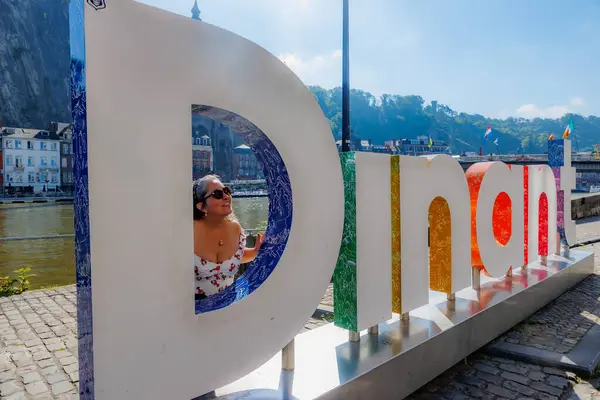 Dinant, Namur, Wallonia, Belçika. 6 Haziran 2024. Yetişkin bir kadın D harfinin başında duruyor. Dinant, binalar, köprü, nehir kenarı, sisli arka plan, gri saç, dekolte bluz ve güneş gözlüğü, güneşli bir gün.