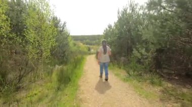 Yolda yürüyen yaşlı kadın yürüyüşçünün görüntüsü, mikrofon elinde, kameraya geri dönüş, arka planda yapraklı ağaçlar, Hoge Kempen Ulusal Parkı 'nda Mechelse Heide, A, Belçika' da güneşli bir gün.