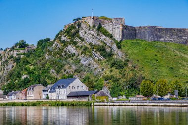 Arka planda mavi gökyüzüne karşı Charlemont kalesi, Givet şehrinin şehir manzarası, ön planda binalar ve Meuse nehri, kalın taş duvarlar, Ardennes departmanında güneşli bir gün, Fransa