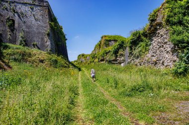 Arka planda mavi gökyüzüne karşı Charlemont Kalesi 'ndeki devasa taş duvarlar, kameraya geri dönen kadın turist, Givet, Ardennes, Fransa' da güneşli bir yaz günü