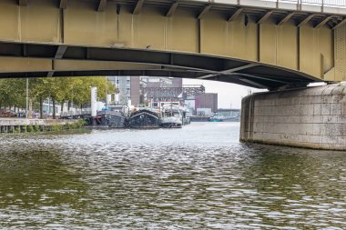 Köprü tabanındaki çelik yapılar, Meuse nehri boyunca rıhtıma demirlemiş gemiler ve bulanık arka planda fabrikalar, Hollanda, Güney Limburg 'daki Maastricht' te bulutlu bir gün.