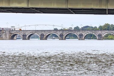 Meuse nehri üzerinde Wilhelminabrug köprüsünün altından geçen Sint Servaasbrug köprüsü arka planda, sakin su, Güney Limburg Maastricht şehrinde bulutlu bir gün