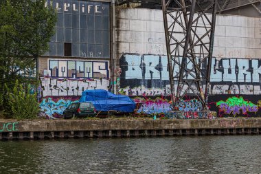 Maastricht, Güney Limburg, Hollanda. 30 Ağustos 2024. Nehir kıyısında kötü bakımlı sokak, eski bir fabrikanın grafiti kaplı duvarı, park edilmiş araba, metal yapı, yeşil ağaçlar, bulutlu bir gün.