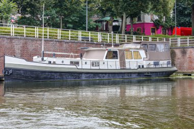 Maastricht şehrinin eski tarihi iç limanının köşesine demir atmış tekne, tuğla duvar, metal korkuluk, arka planda yeşil yapraklar ve evler, Güney Limburg, Hollanda 'da bulutlu bir gün.