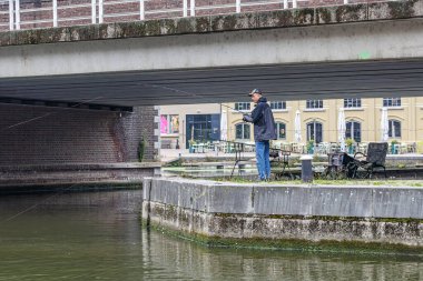 Maastricht, Güney Limburg, Hollanda. 30 Ağustos 2024. Eski tarihi liman, araç köprüsü ve arka plandaki binaların gölgesinde balık tutan genç adam, bulutlu bir gün.