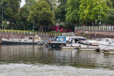 Maastricht şehrinin tarihi iç limanı rıhtımda demirli gemiler, arka planda metal korkuluklar ve bol yapraklı ağaçlarla dolu rampa, Güney Limburg, Hollanda 'da bulutlu bir gün.