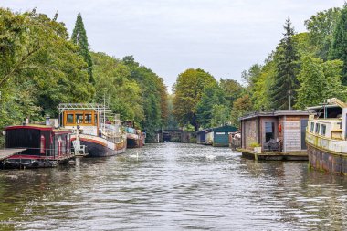 Sahilde yüzen yüzen yüzen yüzen yüzen tekneleri olan gemi kanalı Maastricht şehrinin dışında, ahşap köprü ve arka planda yapraklı ağaçlar, Güney Limburg, Hollanda 'da bulutlu bir gün.