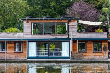 Maastricht şehrinin dışındaki nakliye kanalına demirlemiş iki katlı yüzen ev, aşınmış ahşap duvarlar, büyük pencereler, arka planda yapraklı ağaçlar, Güney Limburg, Hollanda 'da bulutlu bir gün.