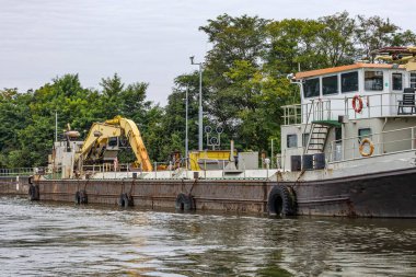 Güvertede kazı yapan eski bir sanayi gemisi, Meuse nehrinin kıyısında küçük bir rıhtımda demirli, arka planda gri gökyüzüne karşı yeşil yapraklı ağaçlar, Güney Limburg, Hollanda 'da bulutlu bir gün.