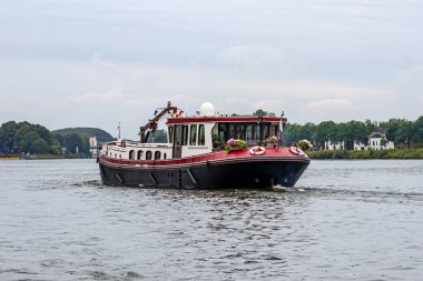 Meuse nehrinde çiçek buketleriyle süslenmiş kırmızı gemi, arka planda tepeler ve yapraklı ağaçlar, Hollanda 'nın güney limanındaki Maastricht şehrinin dışındaki manzara.