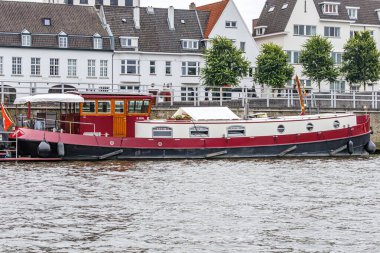 Meuse nehri boyunca rıhtımın rıhtımına demirlemiş kırmızı gemi. Arka planda beyaz duvarlı evler, Hollanda 'nın güney Limburg kentindeki Maastricht şehrinin manzarası.