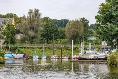 Meuse nehrinin rıhtımında demirli yat ve yelkenli liman, arka planda gri gökyüzüne karşı yapraklı ağaçlar, Maastricht, Güney Limburg, Hollanda 'da bulutlu bir gün.