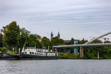 Meuse Nehri, Köprü ve Meryem Ana Bazilikası 'nın rıhtımında demirlemiş bir tekne, arka planda bol yeşil yapraklı ağaçlar, Maastricht, Güney Limburg, Hollanda' da bulutlu bir gün.
