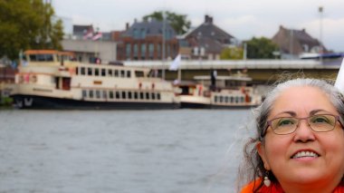 Gözlüklü, gülümseyen olgun bir kadının portresi, Meuse 'nin nehir kıyısındaki iskelede demirlemiş tekneler bulanık arka planda, Maastricht, Güney Limburg, Hollanda' da bulutlu bir günde