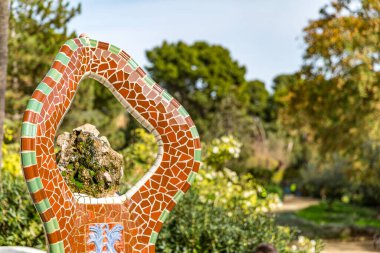 Kahverengi ve yeşil kiremitleri olan içi oyulmuş bir yapı, sürreal tarzı ve şekilleri olan, bulanık arka planda yemyeşil bitki örtüsü, Antoni Gaudi 'nin Parc Guell' i, Barselona, Katalonya 'da güneşli bir gün, İspanya
