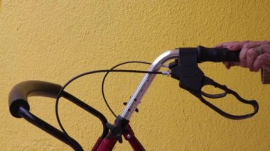 Ayağa kalkıp yürümek için yürüteç tutan yaşlı bir kadının elleri fren kolları, arka planda sarı duvar. Engelli insanların günlük yaşamlarını kolaylaştıracak nesneler kavramı