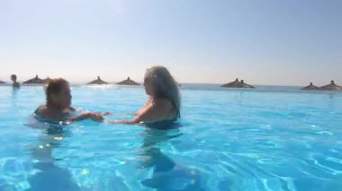 Kızı, yaşlı annesinin açık havuzda yüzmesine yardım ediyor, el ele tutuşuyor, ayak tekmeleriyle hareketli rehabilitasyon yapıyor. İki yetişkin kadın güneşli ve rahatlatıcı bir günün tadını çıkarıyor. Etkin yetişkin yaşam tarzı konsepti