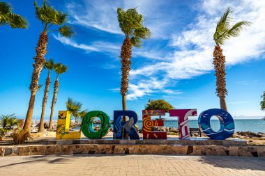 Loreto, Baja California Sur, Meksika. 10 Aralık 2024. Turist gezisi, palmiye ağaçları, arka planda Koyu ve Cortes Denizi 'ne ait renkli harfler. Mavi gökyüzü olan güneşli kış günü.