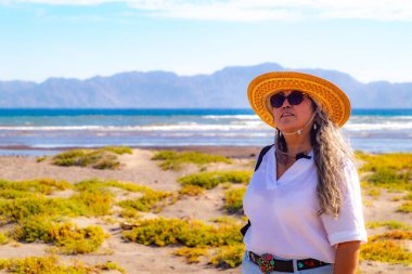 Sahilde başı öne eğik duran, arka planda deniz ve dağlar olan, beyaz bluz ve şapka giyen, kış günü Loreto, Baja California Sur, Meksika 'da bulunan yetişkin bir kadının belden yukarı portresi.