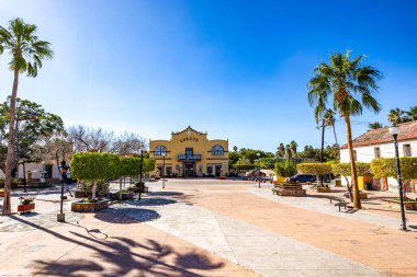 Şehir evleri arasında mavi gökyüzüne karşı sıralanmış yeşil ağaçlarla kaplı ana meydanın geniş merkezi geçidi. Arka planda Hükümet Sarayı. Loreto, Baja California Sur, Meksika 'da güneşli bir kış günü.