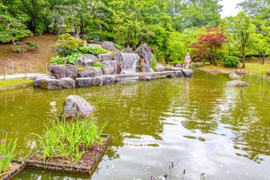 Japon bahçesinde şelaleli gölet, devasa taşların arasından akan su, arka planda yeşil yapraklı ağaçlar, su yüzeyinde yansıma, Hasselt, Belçika 'da bulutlu bahar günü