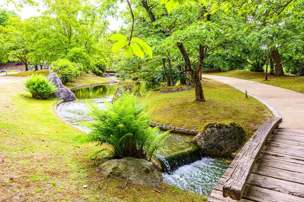 Japon kamu bahçesinin kısmi manzarası. Akan su, yaya yolları, küçük ahşap köprü ve arka planda yemyeşil ağaçlar. Hasselt, Belçika 'da bulutlu bir gün.