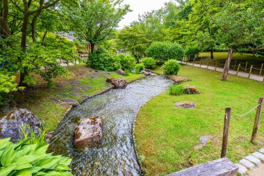 Japon Hasselt bahçesindeki kıvrımlı kanal, arka planda kayboluyor, sakin sular, yaya yolları, yeşil çimenler ve bol yapraklı ağaçlar, Belçika 'da bulutlu bir gün.