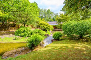 Japon bahçesindeki yapraklı ağaçların manzarası, akan suların kanalı, yeşil çimlerin arasında, yaya yolları, ve arka plandaki doğu evi kısmen manzarası, Hasselt, Belçika 'da güneşli bir gün.