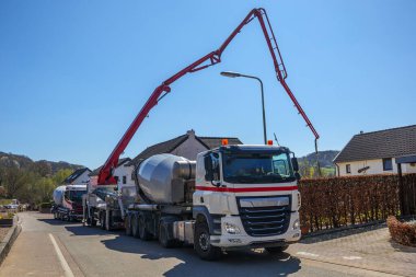 Park halindeki çimento kamyonları ve vinç ile vilayet caddesinde beton boşaltma hortumu, arka plandaki inşaat alanına çimento, Gulpen, Güney Limburg, Hollanda 'da güneşli bir günde