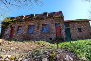 Tepedeki eski, terk edilmiş, harap olmuş bir ev mavi gökyüzüne karşı, yıpranmış tuğla duvarlar, kırık pencereli, metal ve ahşap kapılı çatı, Gulpen, Güney Limburg, Hollanda 'da güneşli bir gün.
