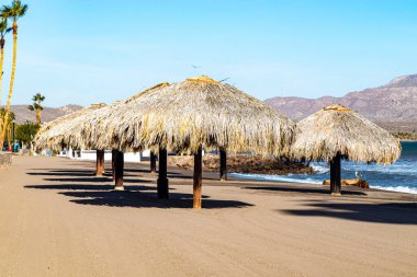 Sahilde bir grup palapanın bulunduğu sahil manzarası, kumsal kumları, kıyıya vuran dalgalar, arka planda dağlar ve mavi gökyüzü, Loreto, Baja California Sur, Meksika 'da güneşli bir bahar günü