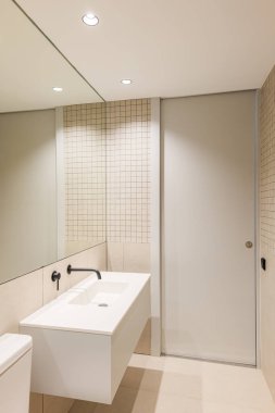 Dar minimalist banyonun dikey görüntüsü. Büyük ayna ve siyah musluklu beyaz lavabo.