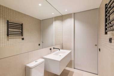 Modern banyo, büyük ayna, bej fayanslar, siyah radyatör ve beyaz lavabo..