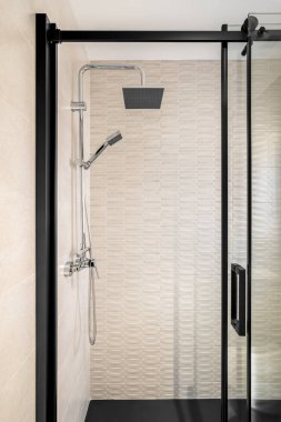 Yağmur başlıklı modern fayanslı banyo, elde tutulan duş ve siyah çerçeveli cam kapı.