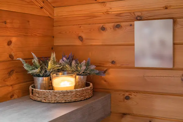Burning candle in glass candlestick with plants as a decor in a bathroom with wooden walls and blurred picture.