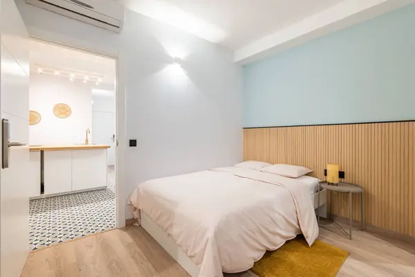 Bright room with a double bed, a bedside table and a terry yellow rug. Simple and minimalistic bedroom interior design in light pastel colors. The doorway reveals a kitchen with modular furniture